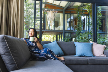 Happy asian woman on couch with mug of coffee thinking at home