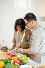 A happy Asian wife is chopping a sweet pepper while her husband is helping her in the kitchen.