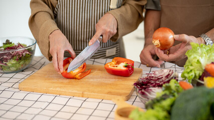 A happy Asian wife is chopping a sweet pepper while her husband is helping her in the kitchen.