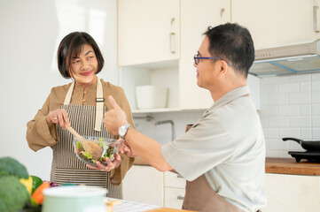 A happy adult Asian husband is showing his thumb up to his wife while enjoying cooking in the kitchen together.