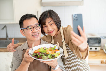 A lovely Asian couple taking pictures or selfies with their food in the kitchen together.
