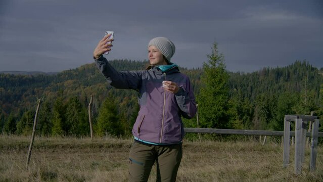 Female hiker takes selfie using mobile phone standing in front of beautiful scenery on the hill. Caucasian woman holds cup of tea and looks at smartphone. Tourist during trip or trek in the mountains.