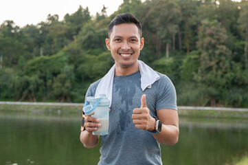 A happy, strong Asian man in sportswear is drinking a protein shake after a long run outdoors.