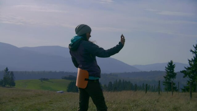 Caucasian female tourist takes selfie in front of beautiful scenery standing on top of the hill. Adult woman looks at photos on her mobile phone. Hiker during expedition or hiking trip in mountains.