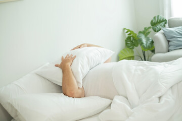 Young over weight man in bed, covering face with pillow, ignored alarm clock. Lazy day in morning on bed.