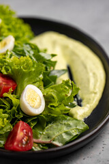 Fresh green salad with quail eggs, cherry tomatoes and guacamole in black plate on grey background