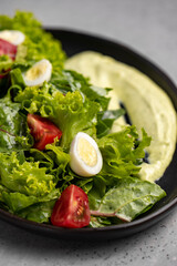 Fresh green salad with quail eggs, cherry tomatoes and guacamole in black plate on grey background