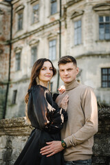 Luxury couple near old Pidhirtsi Castle, Lviv, Ukraine. Woman in black long dress hugs man stand on stairs near ancient palace at sunset. Female embrace male in street. Stylish bride hug groom outdoor