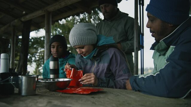 Adult African American man opens doy pack for his wife. Multiethnic tourist family eat hiking food together. Group of travelers or hikers rest in gazebo after expedition or trek in the mountains.