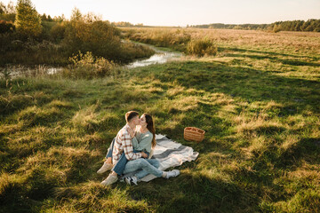 Couple sitting on blanket, hugging and kissing in grass in field at sunset. Young woman, man walking spending time together in nature. Family holiday outdoors. Female embrace male on picnic. Top view