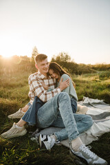 Naklejka na ściany i meble Happy young woman and man, walking spending time together in nature. Couple sitting on blanket, hugging in grass in field at sunset. Concept of family holiday outdoors. Female embrace male on picnic.