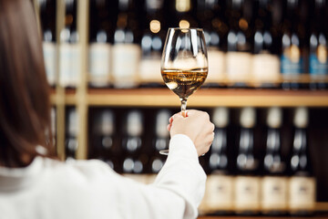 Women hand holds a glass filled with white wine against the background of shelves with various bottles of alcoholic beverages, close up photo. - obrazy, fototapety, plakaty
