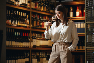 Professional woman sommelier smells white wine from glass, standing in cellar against shelf various alcoholic beverage background.