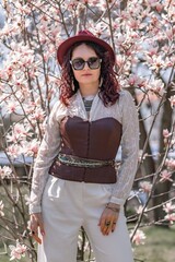 Magnolia park woman. Stylish woman in a hat stands near the magnolia bush in the park. Dressed in white corset pants and posing for the camera.