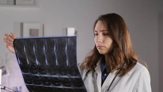 A young female doctor in a white coat holds an X-ray of the bones of the spine and examines it. Medicine and treatment of protrusion and fractures