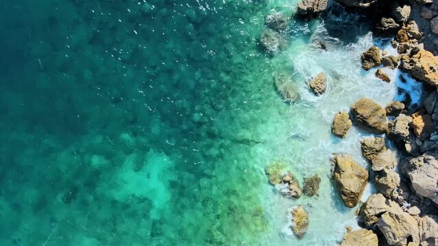 Clear ocean water shimmers as it crashes on rocky coast, nature background drone top down