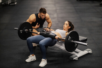 A fit sportswoman is doing workouts for core and abs with barbell in a gym with her trainer. - obrazy, fototapety, plakaty