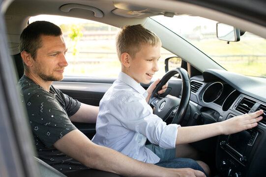 Dad And Happy Son In The Front Seat Of A Car Driving A Car. The Boy Holds The Steering Wheel.