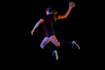 Fototapeta na wymiar Fit young woman engaged in intense handball training, perfecting her throwing and catching abilities against black background in neon light, filter. Concept of sport, hobby, dynamic, championship. Ad