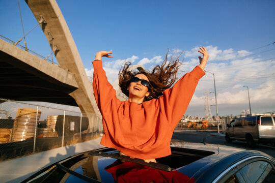An exuberant emotional young woman with excitement as she leans out of the car sunroof with outstretched arms on the city street.