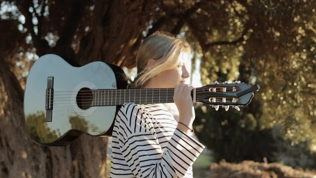 a hipster girl of light appearance with a piercing in her nose stands in the park holding a black guitar on her shoulder, the video was shot around a teenager.