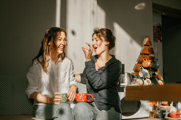 Cozy morning: girls friends talking over a cup of coffee in a bright kitchen Two women spend time peacefully on a sunny day with mugs of tea