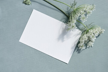 Beautiful white blooming flowers and blank paper card mockup on light blue textured cloth background. Aesthetic floral greeting postcard for Mothers day, Valentines day, wedding invitation template