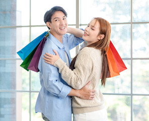 Asian young handsome male boyfriend standing smiling hugging cuddling with beautiful female girlfriend holding paper shopping bags together in romantic decorated living room celebrating valentine day