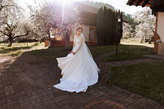 The bride has her shoulders turned to the camera, walking along a stone path. A long train. Magnificent dress with long sleeves, open bust. Spring wedding