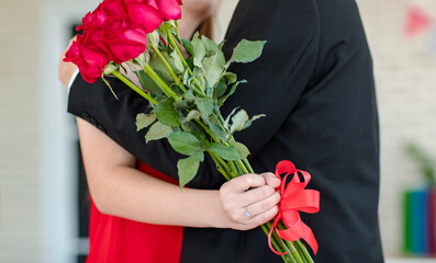 Closeup shot of beautiful red roses flower bouquet in hand of unrecognizable female girlfriend in red dress standing hugging cuddling together kissing with unknown male boyfriend in formal black suit