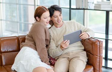 Asian young handsome male boyfriend and beautiful female girlfriend sitting smiling on sofa using tablet computer shopping online via internet together in living room at home