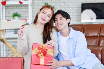 Asian young beautiful female girlfriend sitting smiling opening unboxing red present gift wrapped box on cozy sofa from handsome male boyfriend in romantic decorated living room on valentine day