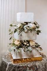 A large luxurious multi-tiered wedding cake is decorated with fresh flowers of white roses and eucalyptus leaves in the banquet hall. Wedding dessert under the evening light. Wedding decor.