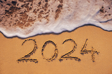 Happy New Year 2024 text written on the beach sand and ocean wave washing the sea shore - obrazy, fototapety, plakaty