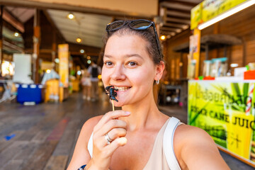 Young woman tourist tasting fried scorpion on a skewer bought in floating open air market on the...