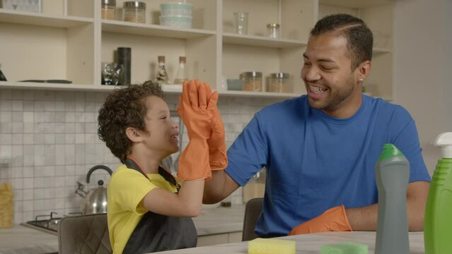 Happy positive school age black boy and cheerful father in protective gloves giving high five gesture before spring cleaning , showing readiness and togetherness in domestic kitchen.