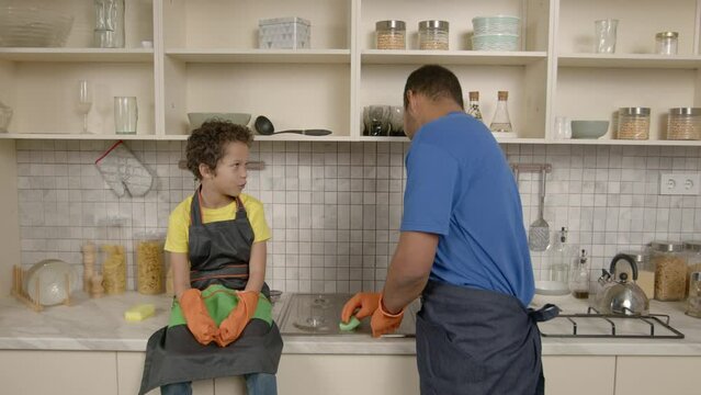 Positive cute preadolescent son in apron and protective gloves sitting on kitchen countertop , talking and sharing with cheerful black father, doing housework together and cleaning kitchen stove .