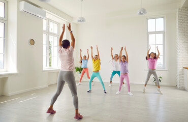 Group of children with help of trainer practice energetic dance moves in choreography class. Young female choreographer shows preteen children dance elements in bright hall of dance studio. - obrazy, fototapety, plakaty