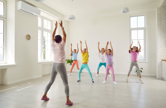 Group of children with help of trainer practice energetic dance moves in choreography class. Young female choreographer shows preteen children dance elements in bright hall of dance studio. - Powered by Adobe
