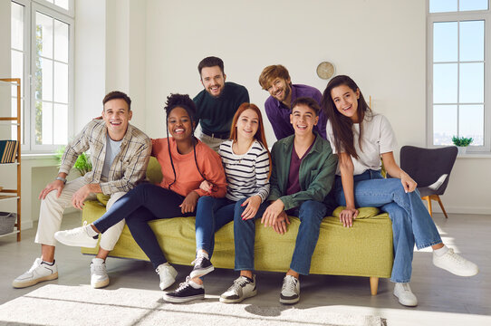 Portrait of a group of happy young diverse friends men and women sitting on sofa in the living room at home looking cheerful at camera enjoying meeting. Friendship, party and home leisure concept.