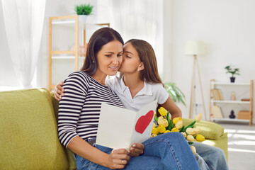 Happy daughter showing love to mom with a surprise card and a beautiful bouquet of flowers on mother day at home. They convey the family comfort, creating an togetherness atmosphere filled with joy.