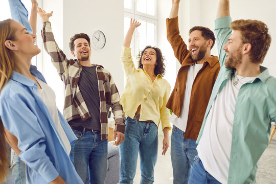 Portrait Of A Group Of Excited Happy Young Friends Students Or Colleagues Having Fun Hanging Together At Home. Men And Women Standing In A Circle Raising Their Hands Up Enjoying Meeting.