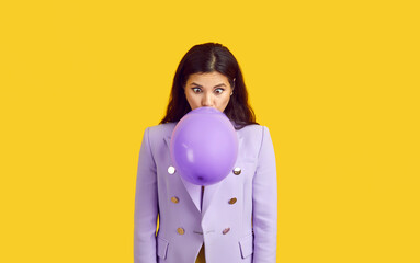 Young woman in trendy jacket blowing up rubber balloon. Beautiful long haired brunette girl in lilac suit isolated on bright yellow background making funny face while inflating purple balloon