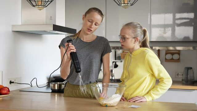 Young beautiful mother explains her daughter how to use electric mixer. Woman melts the butter in the stewpan on electric stove.