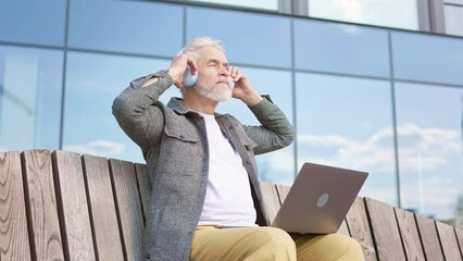 Handsome adult male wearing wireless headphones and immersing himself in music while working on portable laptop outdoors. Serious caucasian man keeping up with time on public area.