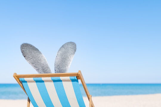 3d rendering of fluffy grey bunny ears protruding from a striped beach chair on a sandy beach with a clear blue ocean in the background. Easter holidays concept.