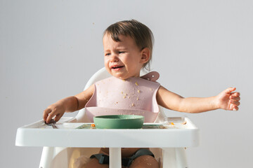 cute crying baby sitting eating lunch in high chair at home on white background, hysterics and tears in a 1 year and 3 month old child - obrazy, fototapety, plakaty