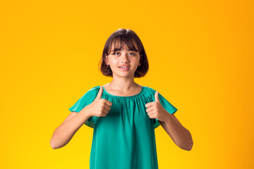 Smiling kid girl showing thumbs up gesture over yellow background. Success concept