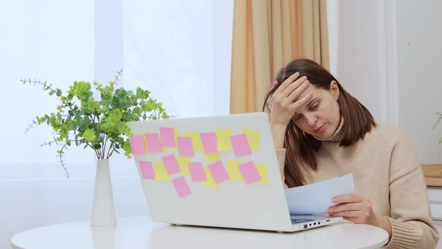 Sad worried Caucasian woman using laptop holding papers paying bills sitting at home in kitchen going over her utility bills thinking about financial problems