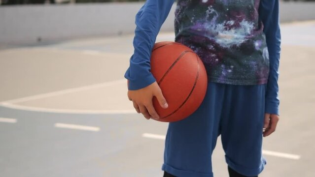 A boy stands on a basketball court and holds a basketball close-up on the ball. A cinematic shot of a teenager standing with a ball in his hand and a basketball basket. High quality FullHD footage